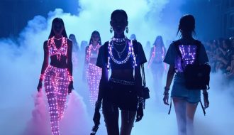 Models on a high-fashion runway wearing vape-inspired accessories, including metallic chain necklaces and iridescent backpacks, set against a background of LED lights that evoke vapor clouds.
