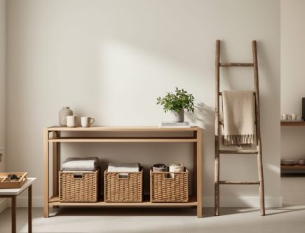 Organized entryway with a key tray, woven baskets under a console table, and a vintage ladder displaying folded throws, with a coffee station suggested in the softly blurred background.