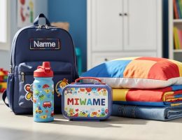 Close-up of neatly arranged children’s personalised everyday essentials—water bottle, embroidered backpack, lunch box, bedding, and folded clothing—in a bright room setting.