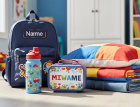 Close-up of neatly arranged children’s personalised everyday essentials—water bottle, embroidered backpack, lunch box, bedding, and folded clothing—in a bright room setting.
